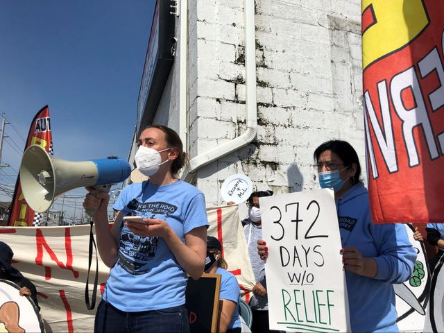 Female protesters wearing masks hold a sign that says "372 Days Without Relief" and a plate that says "Recovery 4 all"